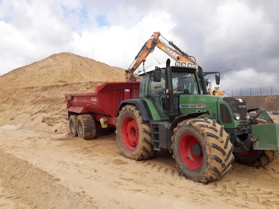 Zielony traktor Fendt 820 z czerwoną przyczepą i koparką CASE, na tle piaskowej góry i pochmurnego nieba. Widoczne ślady opon na piasku.