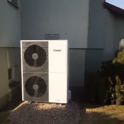 Biała pompa ciepła Vaillant aroTHERM, zainstalowana na żwirowym podłożu przy ścianie budynku w Białaczowie, z widocznymi dwoma wentylatorami i fragmentem ogrodu z tujami w tle.