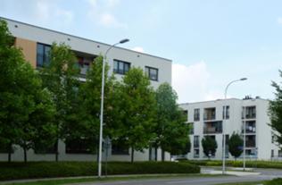 Nowoczesne, białe budynki mieszkalne z balkonami, otoczone zielenią drzew i oświetlone słupami lamp ulicznych, widziane z perspektywy ulicy w słoneczny dzień.