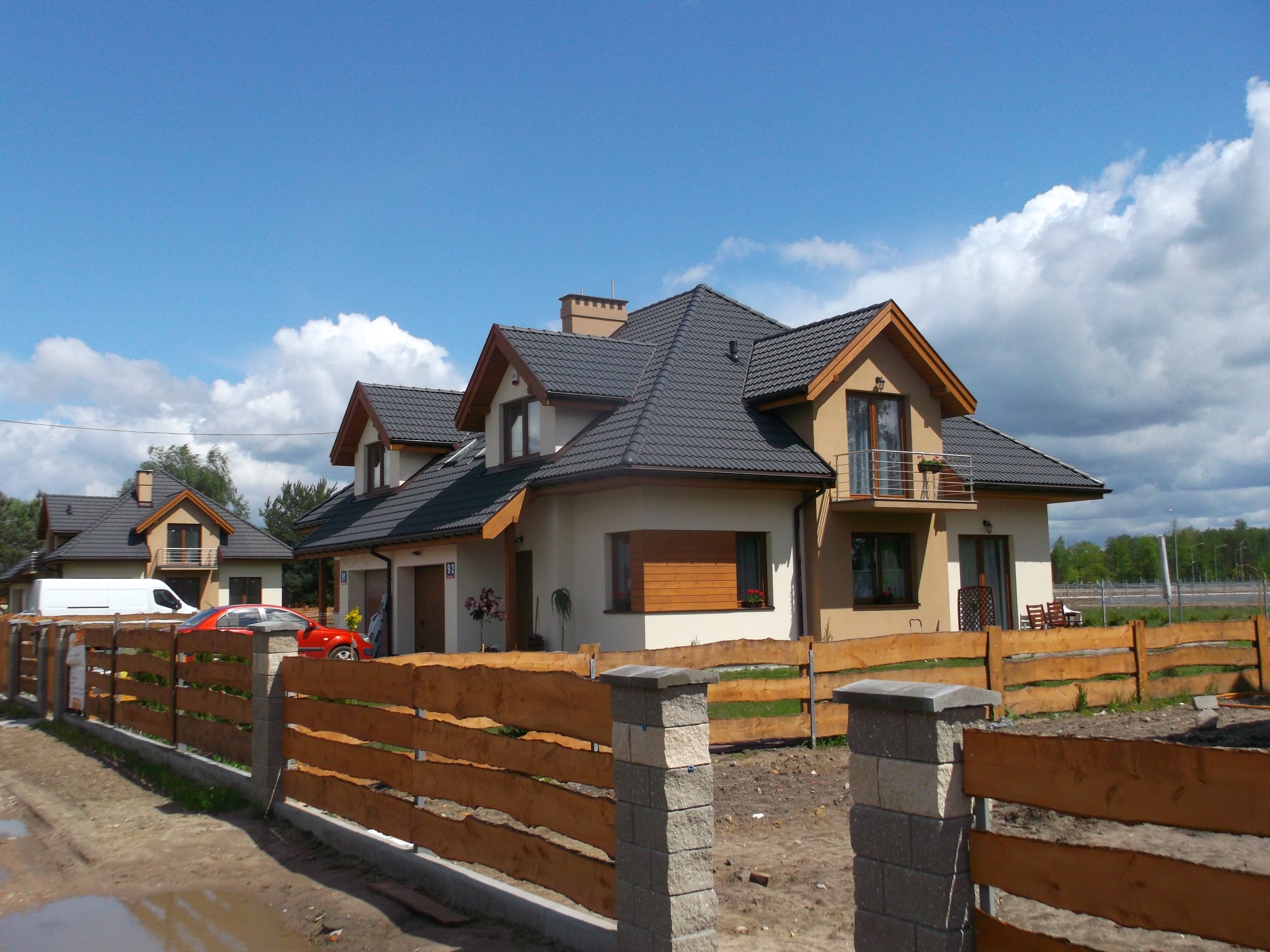 Nowoczesny dom jednorodzinny z ciemną dachówką, drewnianym ogrodzeniem i czerwonym samochodem w tle, widziany w słoneczny dzień.