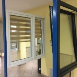 Prezentacja dwóch okien: białe PCV z roletami dzień noc i czarne drewniane, oba uchylone, na tle żółtej ściany.