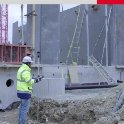 Pracownik w żółtej kurtce i kasku obsługuje urządzenie pomiarowe na placu budowy z surowymi betonowymi ścianami i rusztowaniami w tle.