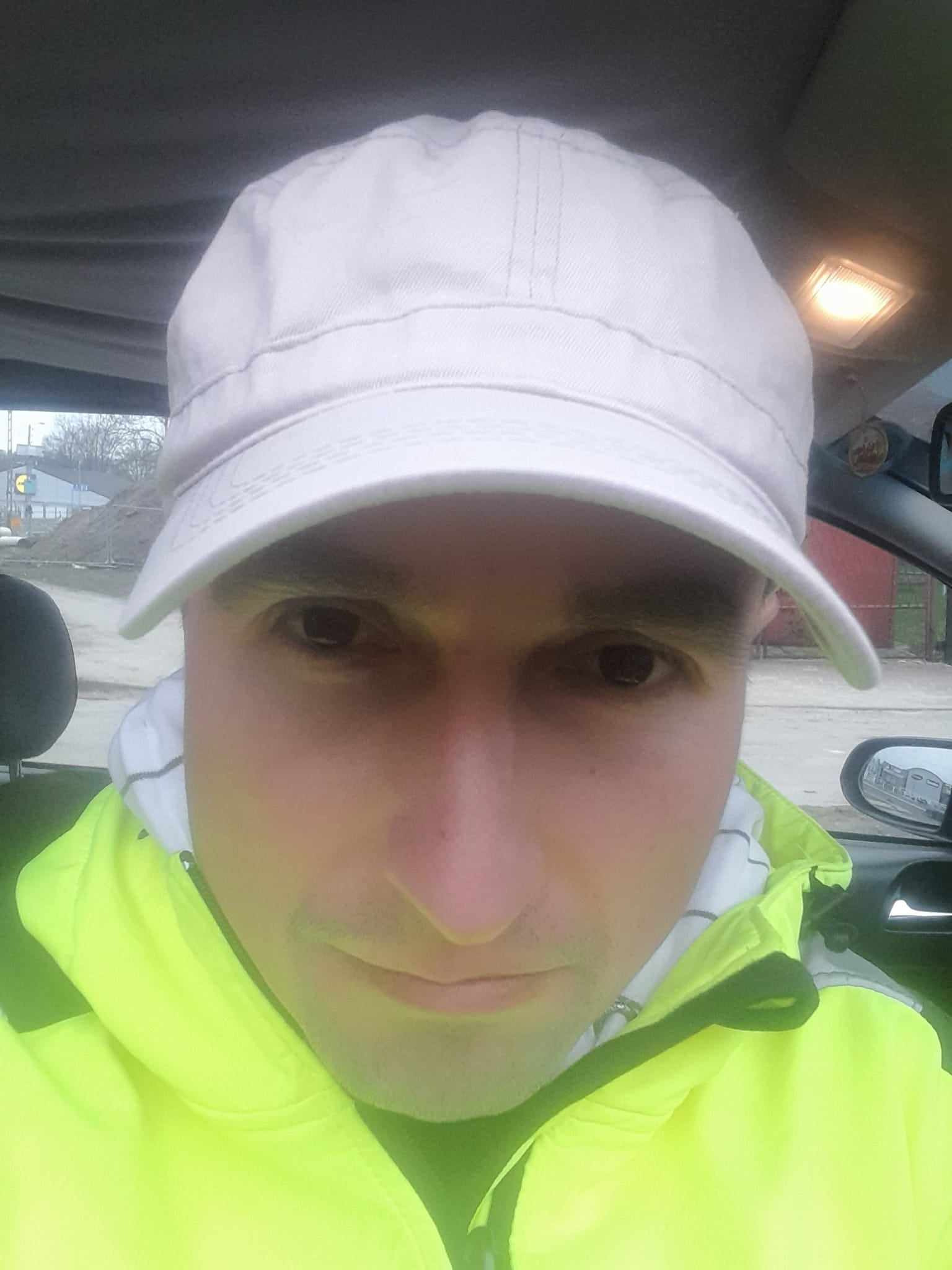 Portret geodety w samochodzie, ubrany w czapkę i jaskrawą kurtkę odblaskową, widok na teren budowy w lusterku bocznym.
