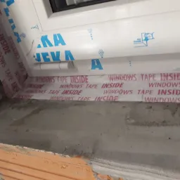 Montaż okna: detal uszczelnienia taśmą okienną 'WINDOWS TAPE INSIDE' na surowej ścianie z cegły, widoczny profil okienny z naklejoną folią ochronną z logo producenta.