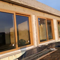 Montaż okien z drewnopodobnymi ramami w surowym budynku z betonu komórkowego, widoczne odbicie krajobrazu w szybach i cień osoby fotografującej.