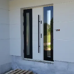 Nowoczesne drzwi wejściowe z czarnymi akcentami i długim uchwytem, jeszcze nie wykończone, widoczne palety pod drzwiami.