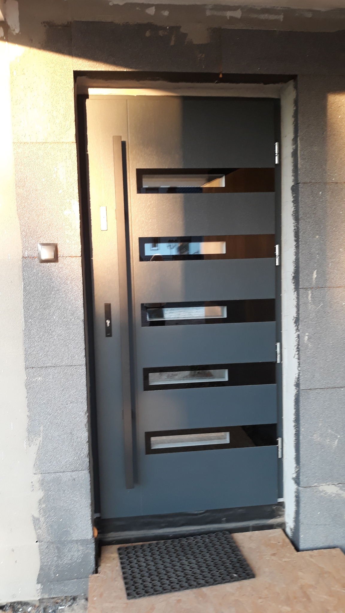 Nowoczesne, szare drzwi wejściowe z poziomymi przeszkleniami, osadzone w surowej, nieotynkowanej ścianie z bloczków betonowych; widoczna wycieraczka przed progiem.