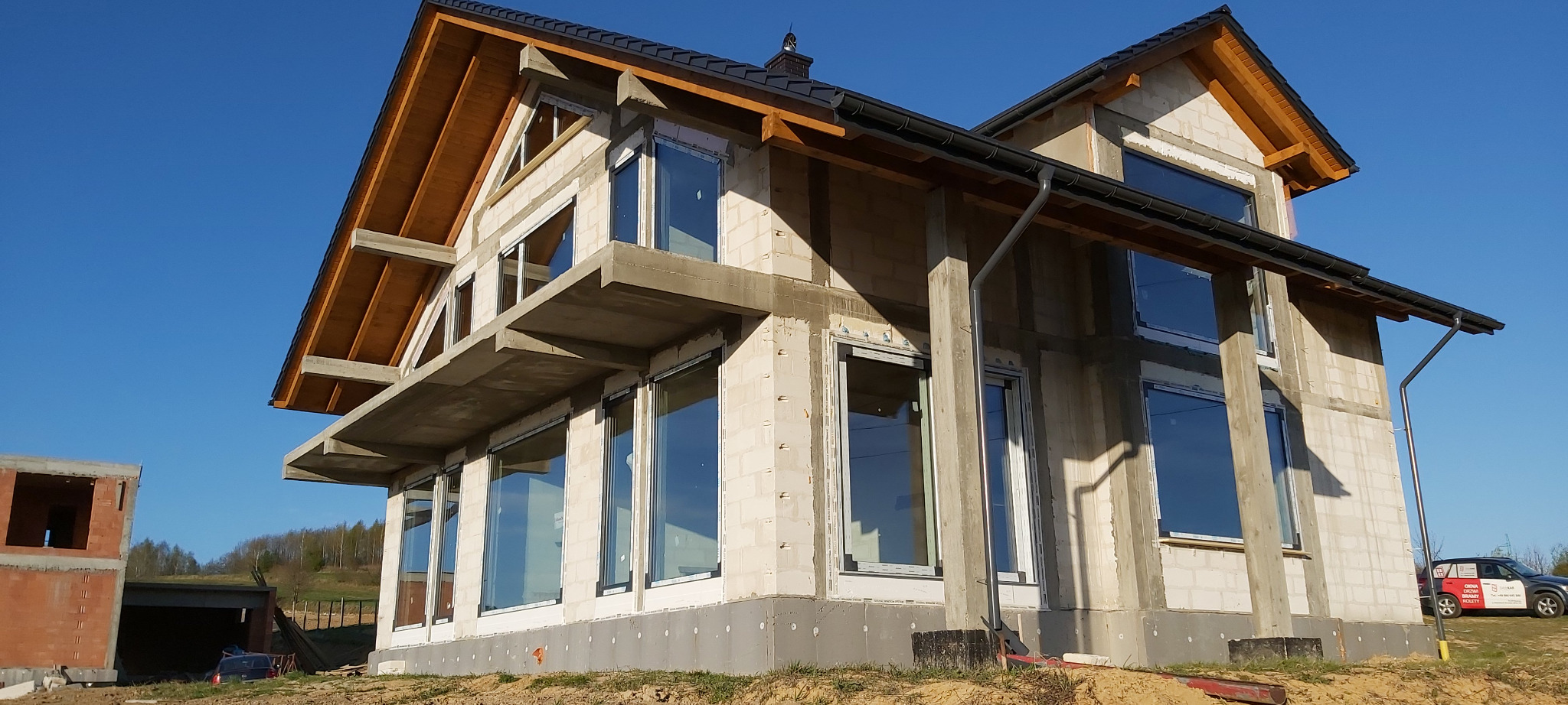 Dom w trakcie budowy z zamontowanymi oknami, widoczne bloczki betonowe i drewniana konstrukcja dachu, na zewnątrz budynku zaparkowany samochód.
