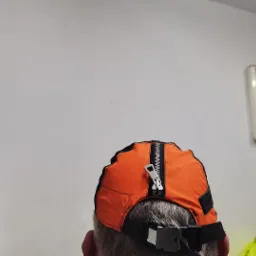 Pomarańczowa czapka z zamkiem błyskawicznym na górze, widok z tyłu głowy, zapięcie z klamrą na pasku regulacyjnym.