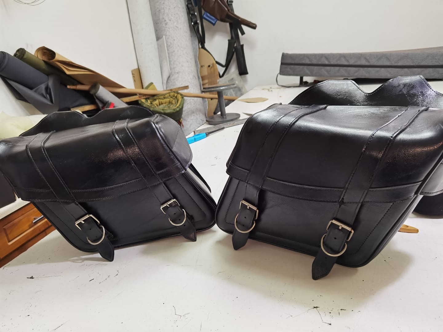 Dwie czarne, skórzane torby motocyklowe z metalowymi sprzączkami, leżące na białym stole w warsztacie, widoczne materiały i narzędzia w tle.