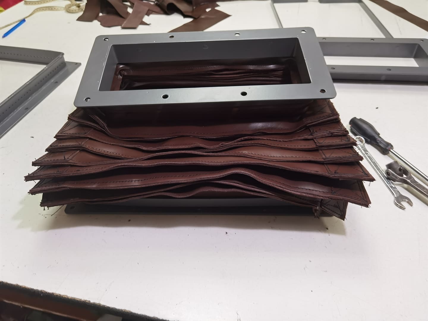 Brązowy, skórzany mieszek harmonijkowy z ramą montażową, widoczne narzędzia na stole warsztatowym.