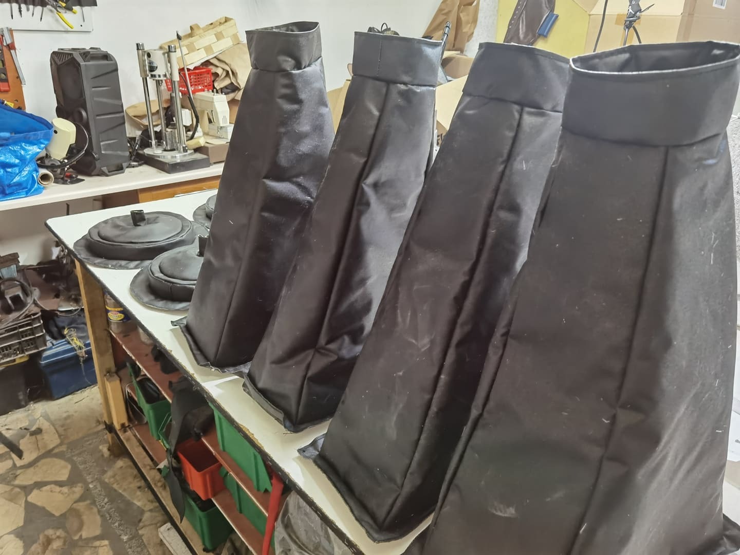 Cztery czarne, materiałowe pokrowce w kształcie ściętego stożka z usztywnioną górą stoją na stole warsztatowym, w tle widoczne narzędzia i maszyny.