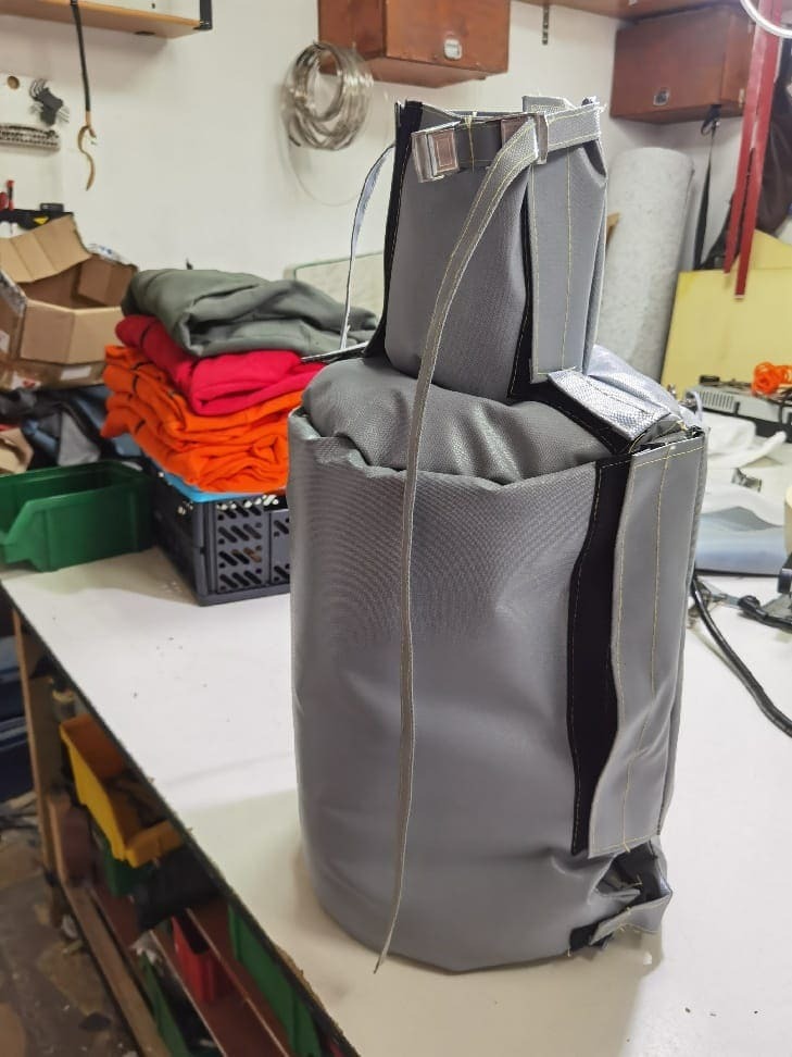 Szary, cylindryczny pokrowiec termoizolacyjny z paskami i klamrami na warsztatowym stole, widoczne stosy kolorowych tkanin w tle.