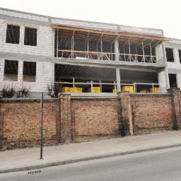 Budowa dwupiętrowego budynku z białych bloczków, widoczna zza ceglanego muru z żółtymi paczkami materiałów budowlanych na drugim piętrze.