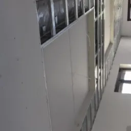 Ściana w trakcie montażu płyty gipsowo-kartonowej na metalowym stelażu, widoczne okna w tle, surowy stan wykończenia wnętrza.