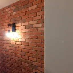 Ściana z czerwonej cegły z czarnym kinkietem oświetlającym fragment pomieszczenia, kontrastująca z gładką, jasną ścianą po prawej stronie kadru.
