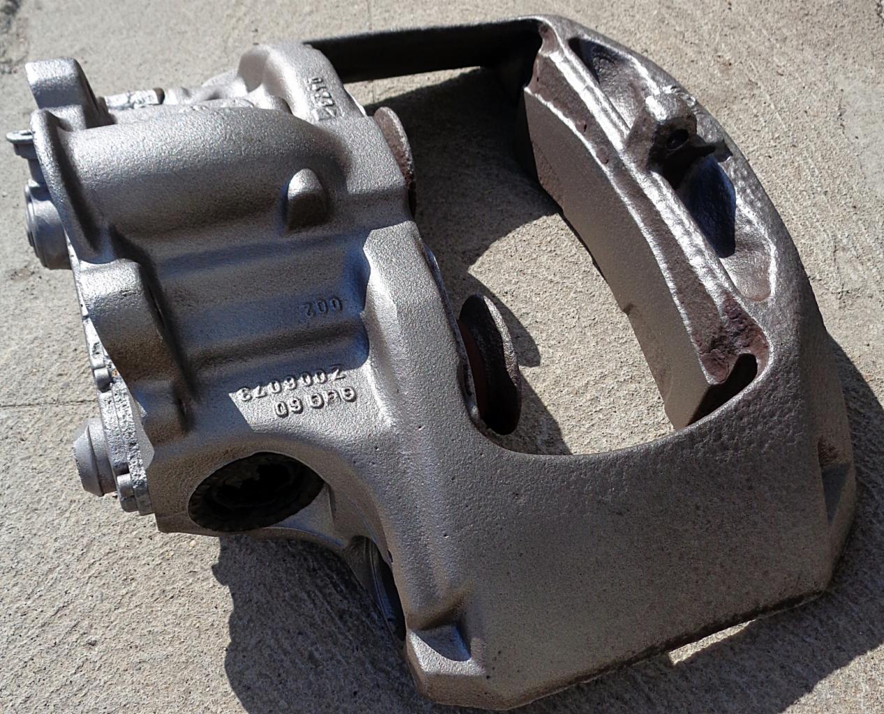 Szary, metalowy zacisk hamulcowy z widocznymi numerami seryjnymi leżący na betonowej powierzchni w mocnym świetle słonecznym.