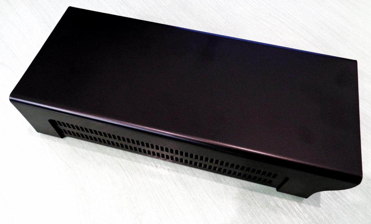 Czarna, metalowa obudowa urządzenia z perforowanymi otworami wentylacyjnymi, leżąca na jasnej powierzchni.