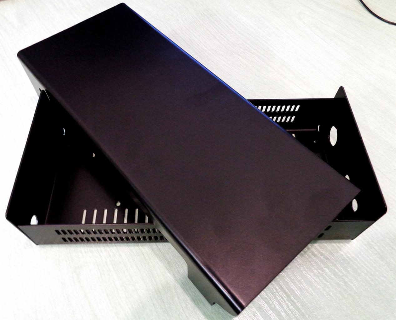 Czarna, metalowa obudowa urządzenia elektronicznego z perforowanymi otworami wentylacyjnymi i zdejmowaną pokrywą, leżąca na jasnym blacie.