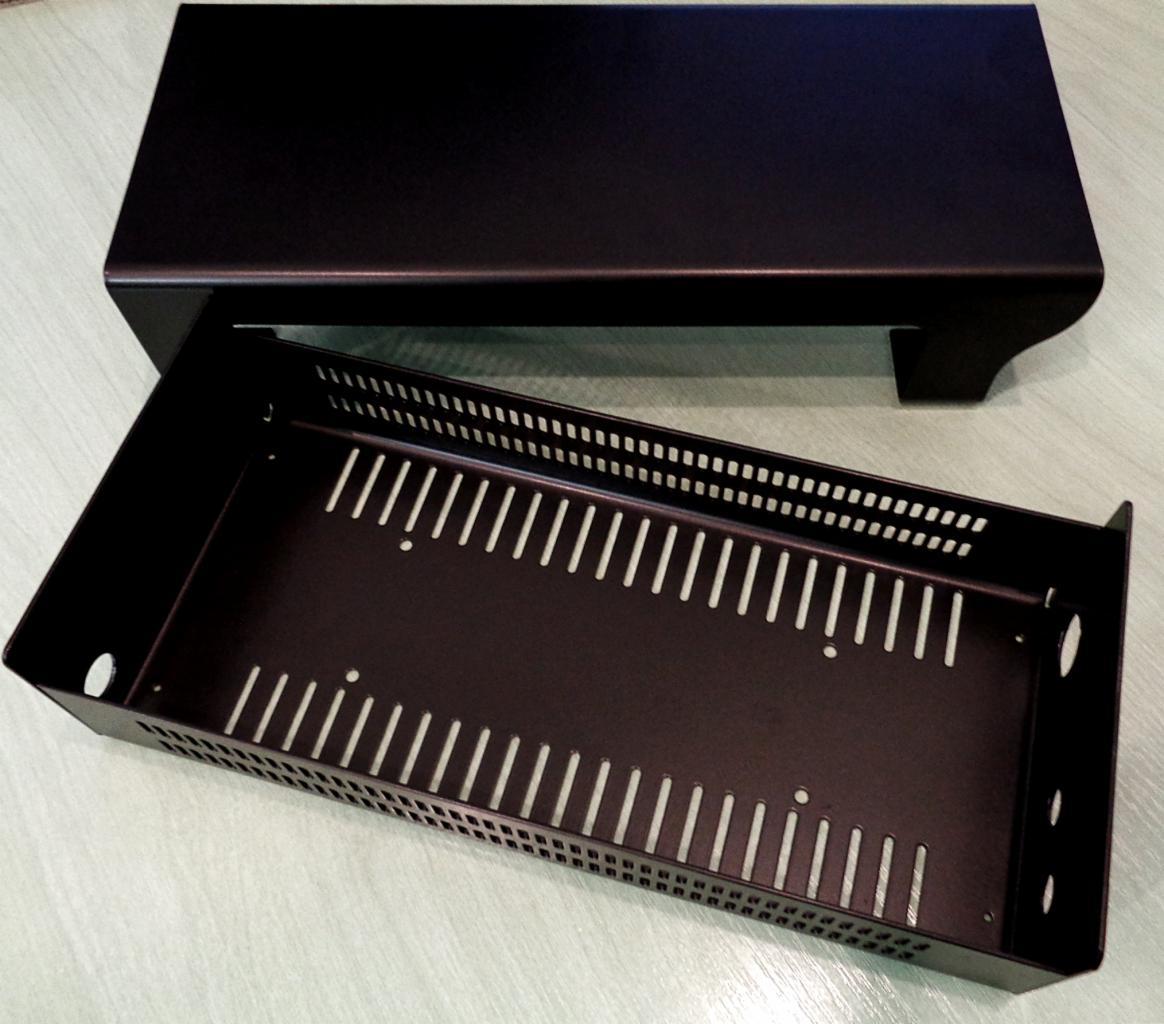 Czarna, metalowa obudowa z perforowanymi bokami i podłużnymi otworami, częściowo wysunięta spod górnej, gładkiej pokrywy.