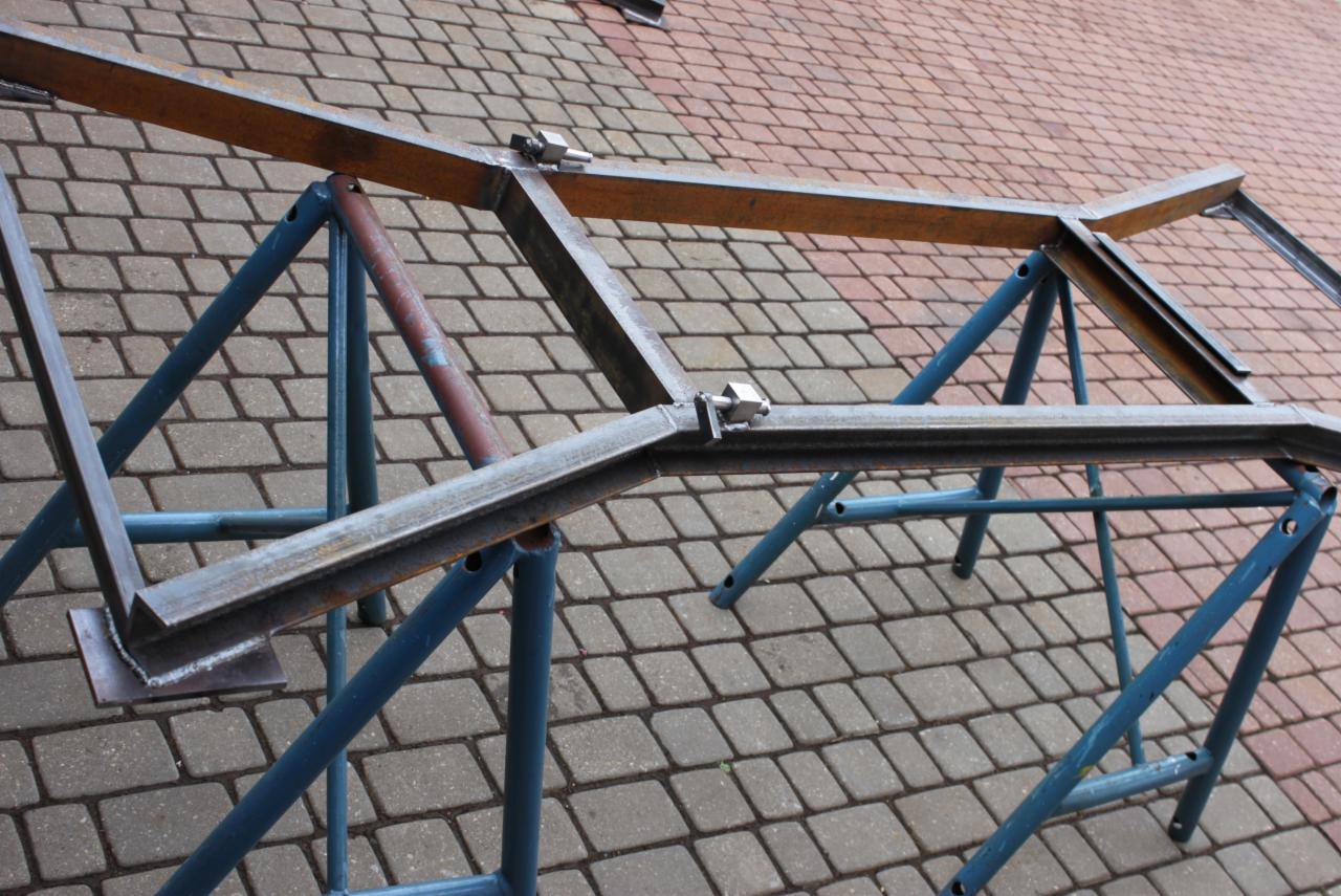 Metalowa konstrukcja spawana z profili, umieszczona na niebieskim stojaku, widoczna na tle kostki brukowej.
