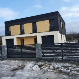 Nowoczesny dom z czarno-biało-beżową elewacją i poziomą, metalową bramą w kolorze grafitowym, na tle zimowego krajobrazu z resztkami śniegu.