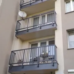 Fasada bloku mieszkalnego z balkonami zabezpieczonymi czarnymi, metalowymi balustradami. Na balkonie na drugim piętrze widoczne krzesła i stolik. Balkon na najniższym piętrze podparty metalowymi...