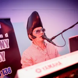 Mężczyzna w paski koszuli i futurystycznym nakryciu głowy śpiewa do mikrofonu, grając na klawiszach Yamaha, obok baner reklamowy zespołu.