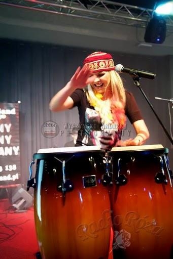 Uśmiechnięta blondynka w czerwonej czapce gra na bębnach conga na scenie, z mikrofonem i kolumnami w tle.