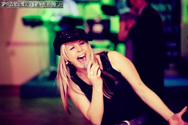 Wokalistka z blond włosami w czarnym kapeluszu i błyszczącej sukience śpiewa do mikrofonu na scenie z zielonym podświetleniem, w tle widoczni goście.