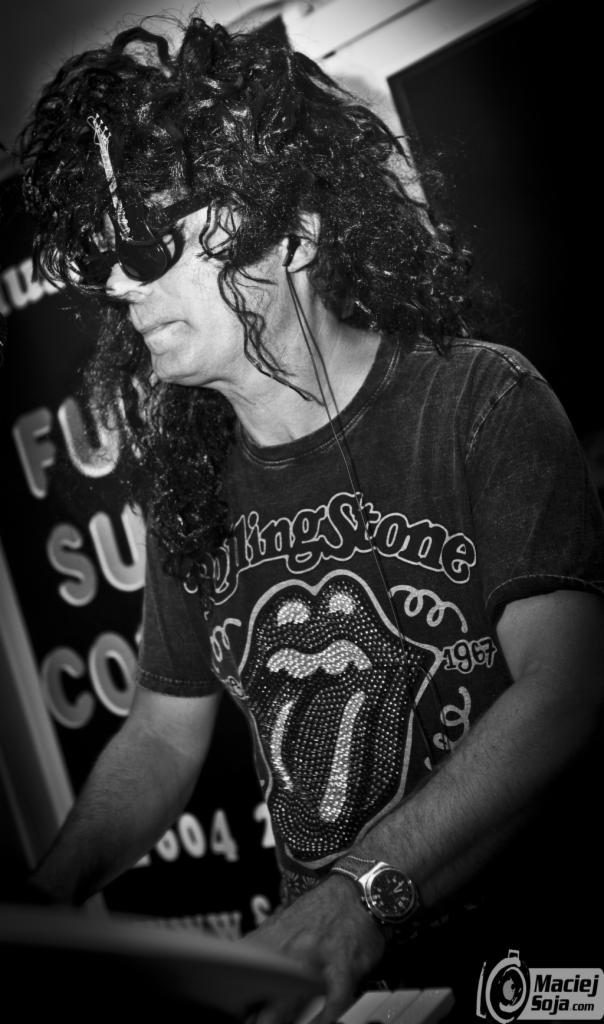 Czarno-białe zdjęcie muzyka z kręconymi włosami w koszulce Rolling Stones grającego na instrumencie klawiszowym, z zegarkiem na ręce i słuchawkami w uszach.