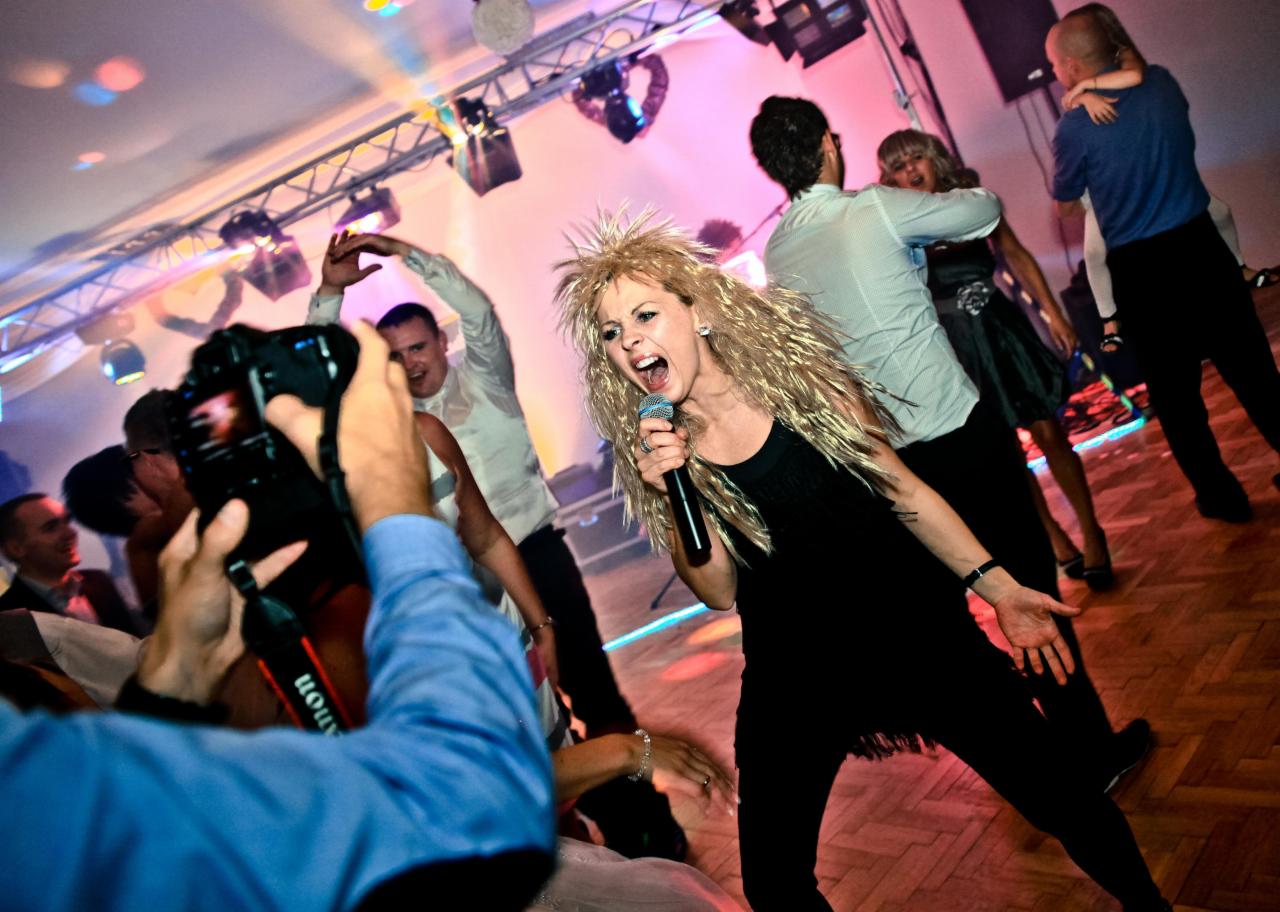 Wokalistka w czarnej sukience z frędzlami i blond peruce śpiewa do mikrofonu na scenie podczas imprezy, w tle tańczący tłum i oświetlenie sceniczne.