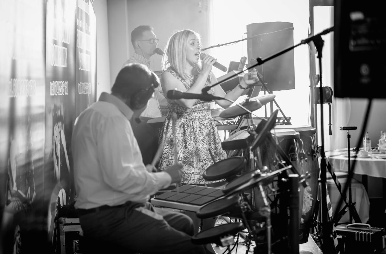 Występujący na scenie zespół: wokalistka z mikrofonem, perkusista grający na elektronicznej perkusji oraz klawiszowiec w tle, ustawieni przed ścianą z plakatem reklamowym.