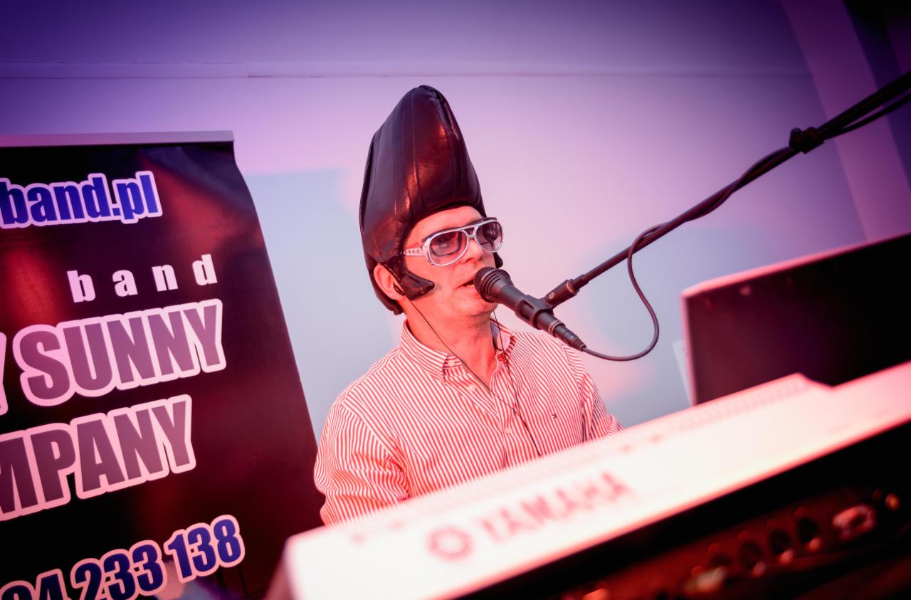 Mężczyzna w paski koszuli i futurystycznym nakryciu głowy śpiewa do mikrofonu, grając na klawiszach Yamaha, obok baner reklamowy zespołu.