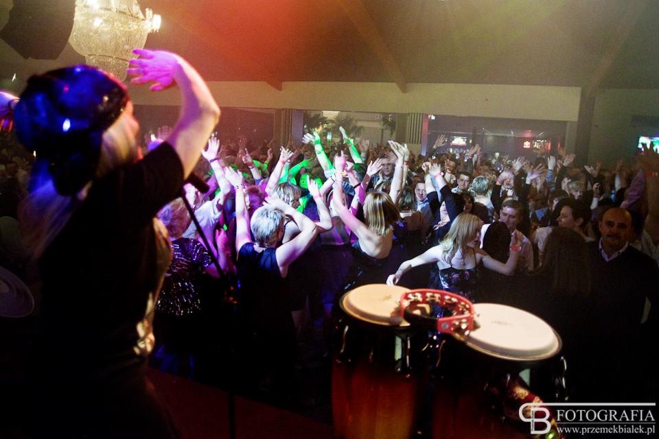 Wokalistka z mikrofonem na scenie, w tle rozbawiony tłum z uniesionymi rękami, bębny conga na pierwszym planie, oświetlenie dyskotekowe.