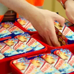 Pracownik w odblaskowej kamizelce wkłada batoniki w kolorowych opakowaniach do czerwonych pojemników podczas procesu konfekcjonowania.