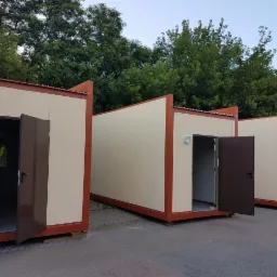 Trzy modułowe kontenery biurowe z otwartymi drzwiami, ustawione na parkingu z widocznymi drzewami w tle.