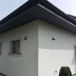 Elewacja domu jednorodzinnego z dwoma anemostatami na ścianie, widoczny fragment dachu z kominem i oknami z luksferami.