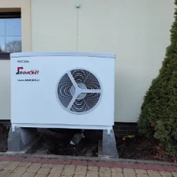 Biała pompa ciepła marki Stiebel Eltron Inmont, zamontowana na betonowych podstawach przy ścianie budynku z oknem, obok zielonego krzewu.