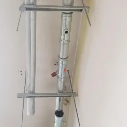 Montaż rur instalacji grzewczej z metalowymi wspornikami w surowym wnętrzu, widoczne otwory w ścianie i podłodze.