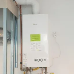 Nowy, biały piec gazowy Beretta zamontowany na białej ścianie, z widocznymi miedzianymi rurami, białym przewodem wentylacyjnym i metalową konstrukcją ściany w tle. Na pierwszym planie fragment...