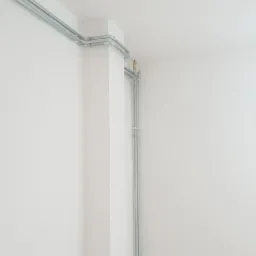 Szare rury instalacyjne poprowadzone wzdłuż ściany i sufitu, widoczne mocowania, białe ściany i sufit, kąt ujęcia od dołu.