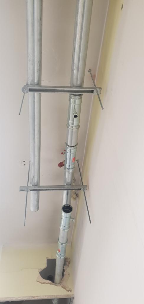 Montaż rur instalacji grzewczej z metalowymi wspornikami w surowym wnętrzu, widoczne otwory w ścianie i podłodze.