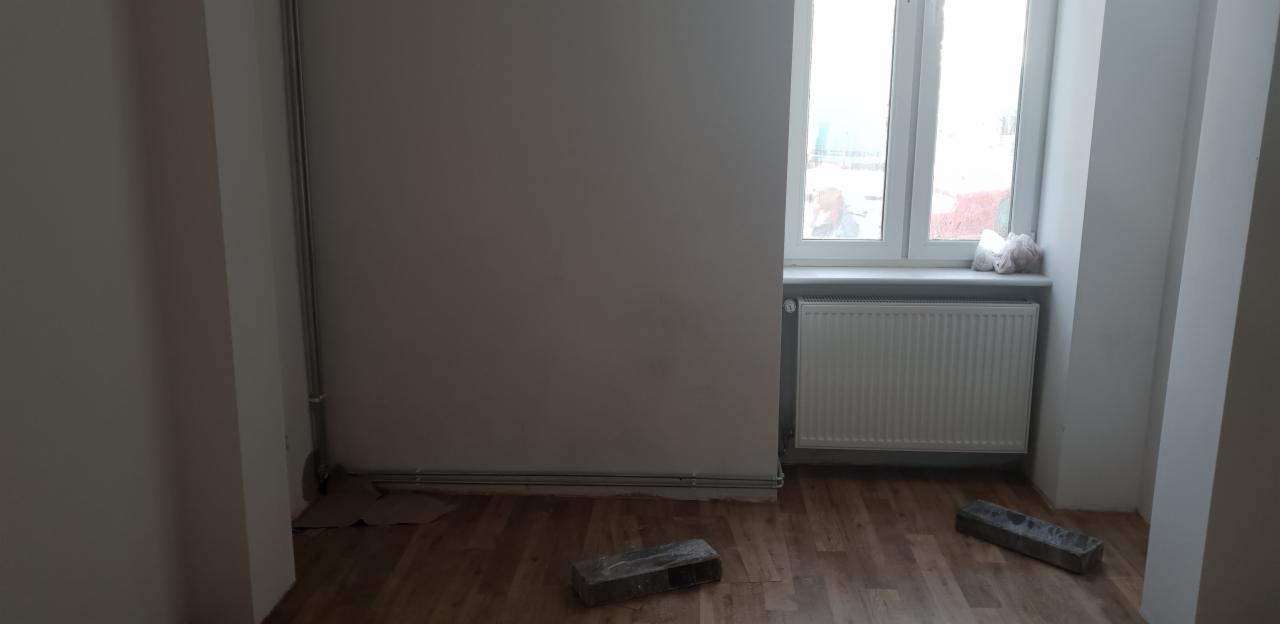 Pusta przestrzeń z grzejnikiem pod oknem, widoczne rury grzewcze przy ścianie i dwa bloczki na podłodze imitującej drewno, przygotowana do dalszych prac wykończeniowych.