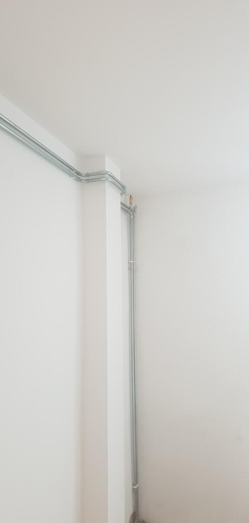 Szare rury instalacyjne poprowadzone wzdłuż ściany i sufitu, widoczne mocowania, białe ściany i sufit, kąt ujęcia od dołu.