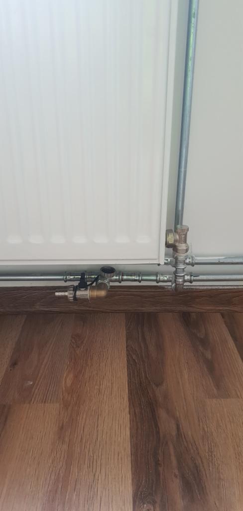 Biały grzejnik panelowy z podłączonymi rurami miedzianymi i zaworami termostatycznymi na tle drewnopodobnej podłogi.