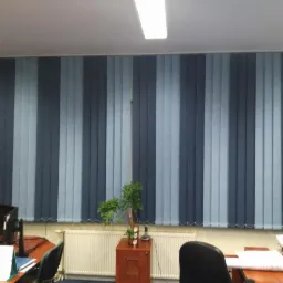 Biuro z pionowymi panelami okiennymi w odcieniach szarości i błękitu, widoczne biurko z komputerem i rośliną doniczkową.