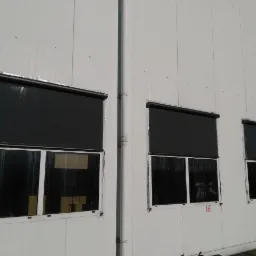 Trzy okna w białej elewacji z zamontowanymi, zasuniętymi, czarnymi roletami zewnętrznymi, zabezpieczone folią ochronną, widoczna kamera monitoringu.