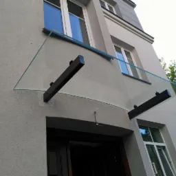 Szklany daszek nad wejściem do budynku, wsparty na dwóch czarnych, prostokątnych wspornikach, widok z dołu.