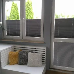 Szare, plisowane osłony okienne zamontowane w oknach i drzwiach balkonowych, widok z wnętrza pokoju, z trzema dekoracyjnymi poduszkami na parapecie.